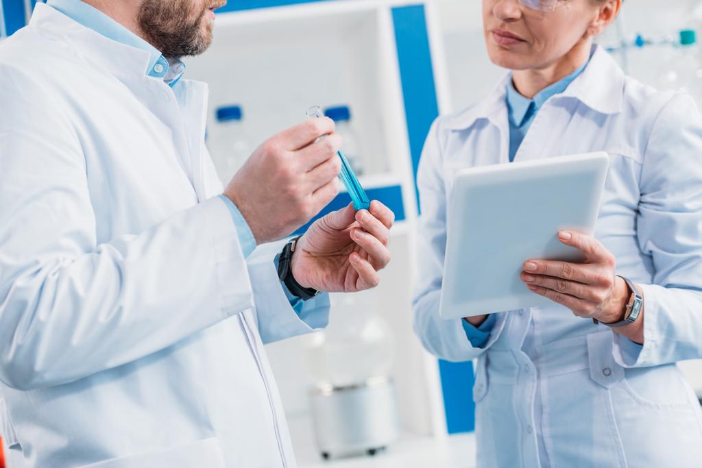 обрезанный снимок ученых в белых халатах с трубкой с реагентом и таблеткой в лаборатории
 - Фото, изображение