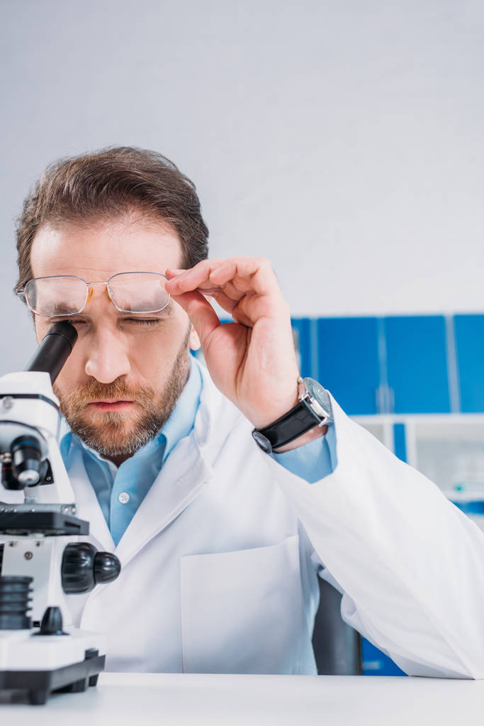 scientist in white coat and eyeglasses looking through microscope on reagent in laboratory - Photo, Image