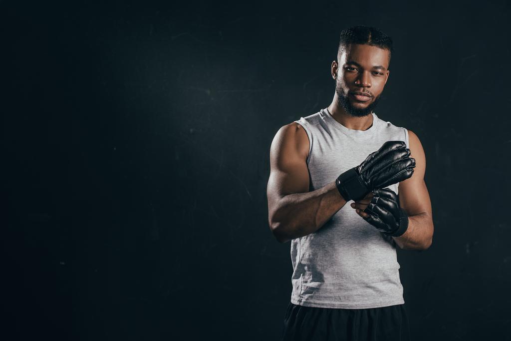 junger afrikanisch-amerikanischer Kickboxer trägt Handschuhe und schaut isoliert auf schwarz in die Kamera - Foto, Bild