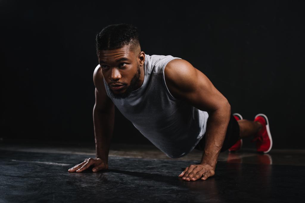 giovane uomo muscoloso africano americano in abbigliamento sportivo facendo flessioni e guardando lontano sul nero
 - Foto, immagini