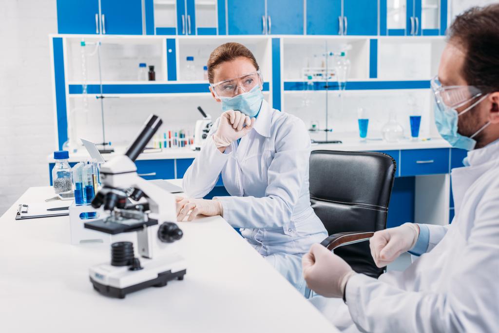 наукові дослідники в медичних масках і окулярах обговорюють на робочому місці з мікроскопом в лабораторії
 - Фото, зображення