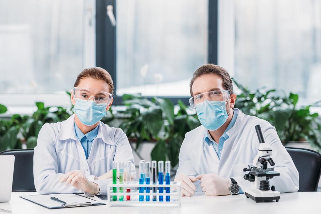 wetenschappelijke onderzoekers in bril en medische maskers zittend op werkplek met reagentia in buizen en Microscoop in lab - Foto, afbeelding