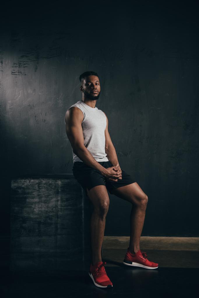 volle lengte weergave van jonge atletische Afro-Amerikaanse man in sportkleding zitten en kijken naar camera op zwart  - Foto, afbeelding