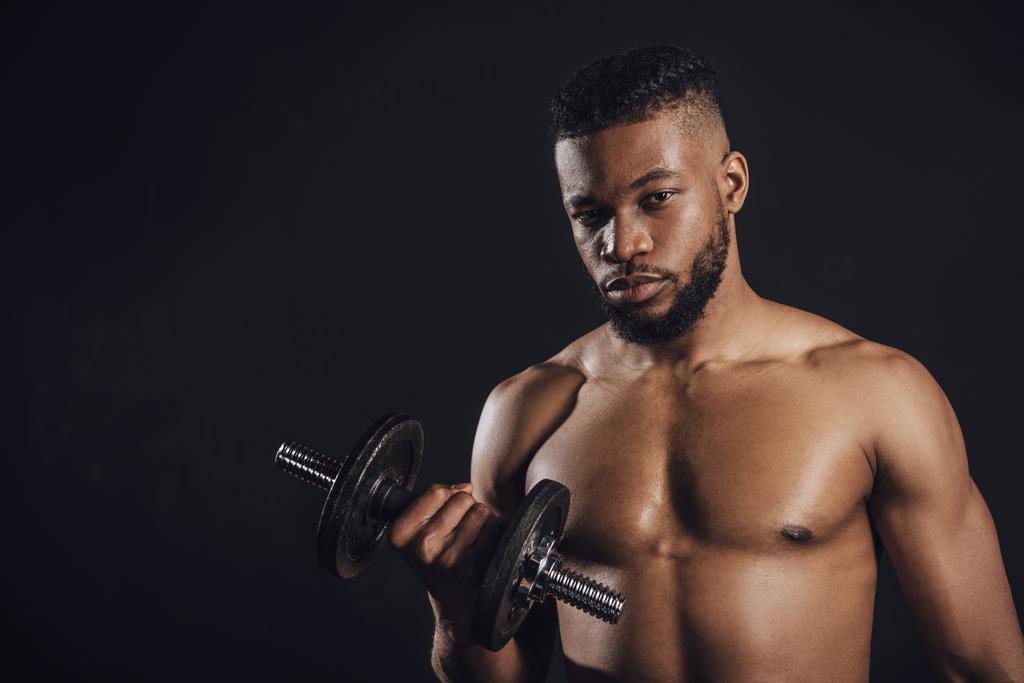 mięśni shirtless african american sportowca szkolenie z hantle i patrząc na kamery na czarnym tle   - Zdjęcie, obraz