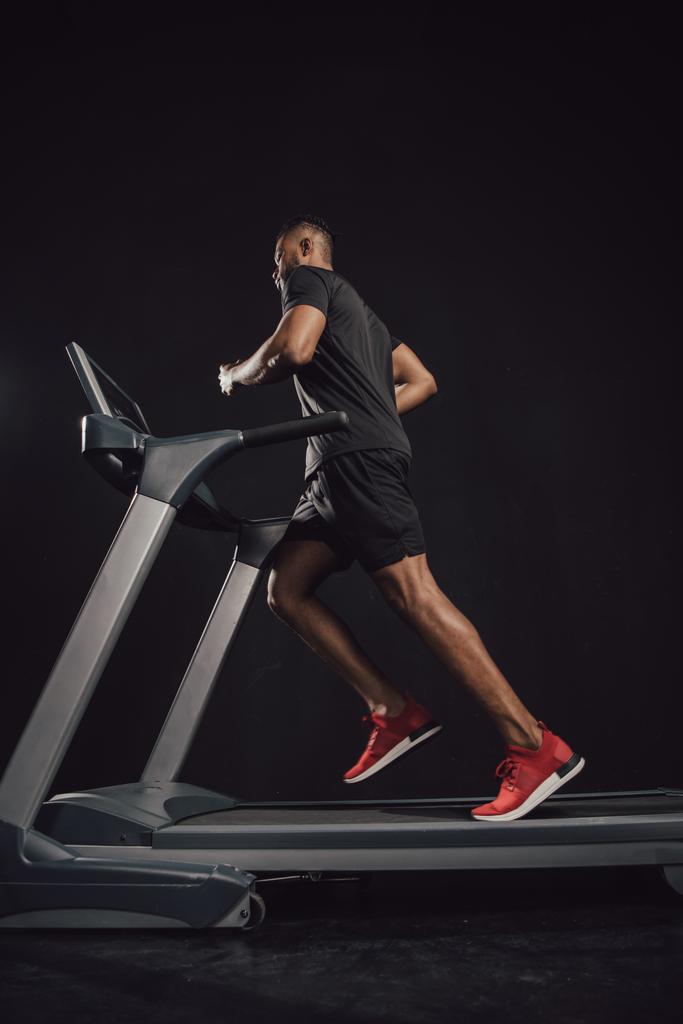 Tiefansicht eines jungen afrikanisch-amerikanischen Sportlers, der auf einem Laufband auf schwarz läuft  - Foto, Bild