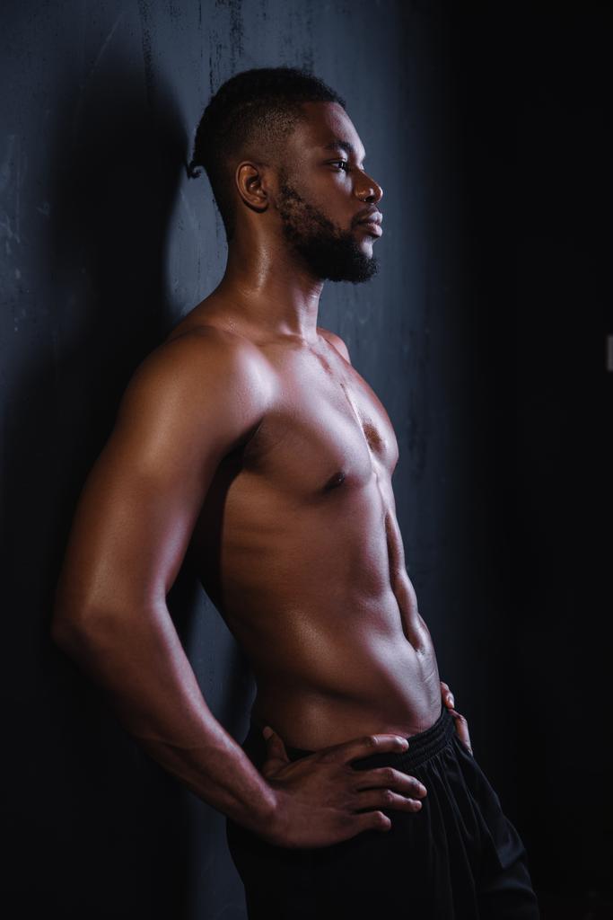 vista lateral del joven afroamericano musculoso sin camisa de pie con las manos en la cintura y mirando hacia otro lado en negro
 - Foto, imagen