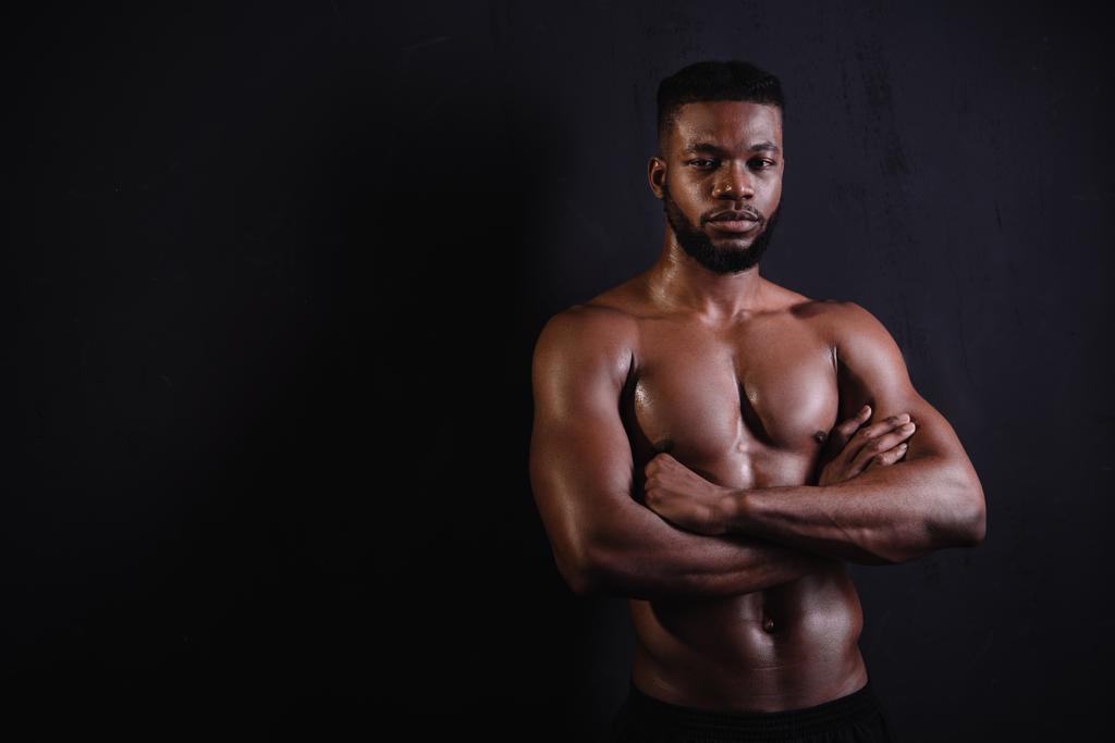 handsome muscular african american man standing with crossed arms and looking at camera on black - Photo, Image