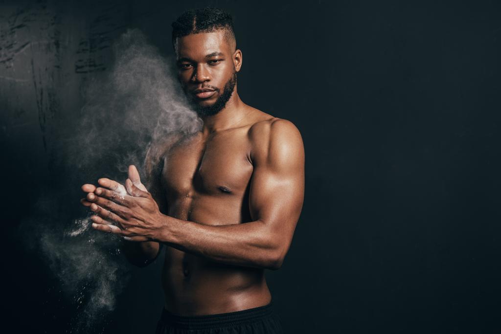νεαρός γυμνός-chested Αφρικανικός Αμερικανός αθλητής εφαρμογή ταλκ και κοιτάζοντας την κάμερα στο μαύρο    - Φωτογραφία, εικόνα