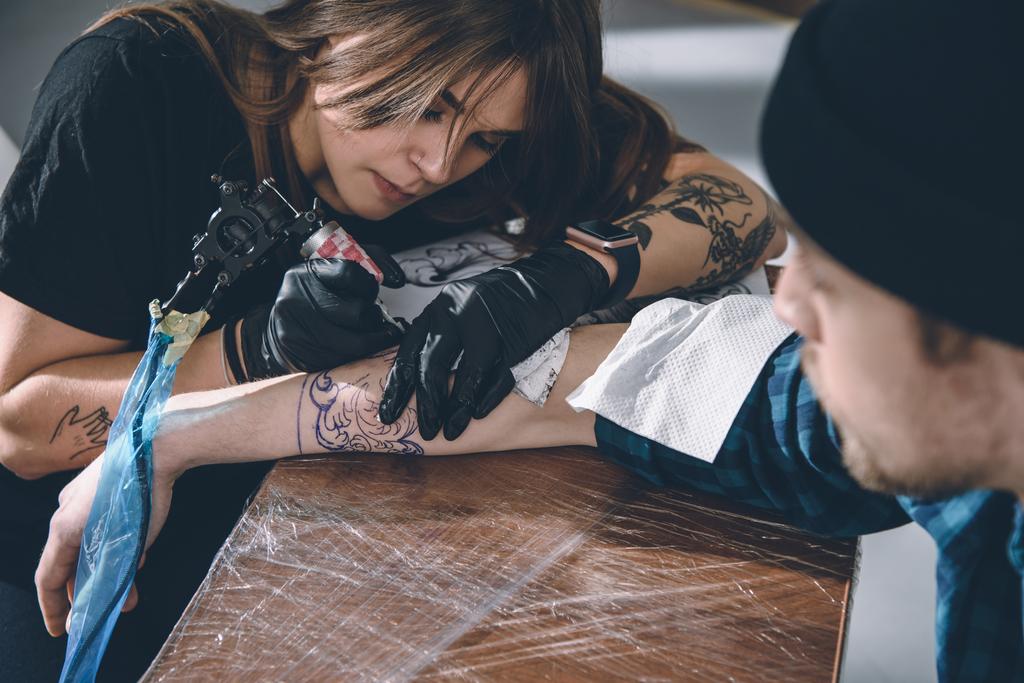 Mestre de tatuagem feminina em luvas trabalhando em peça de braço em estúdio
 - Foto, Imagem
