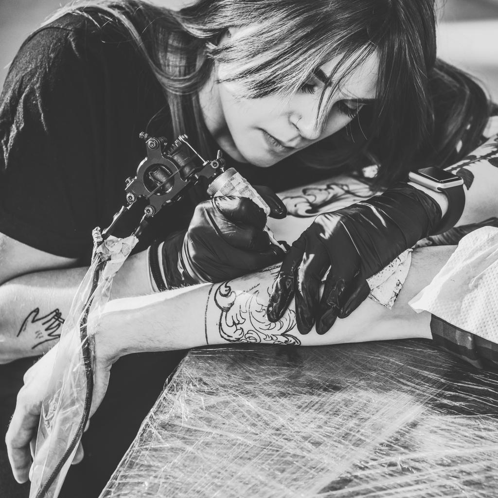 Black and white photo of concentrated female artist in gloves working on arm piece tattoo in studio - Photo, Image