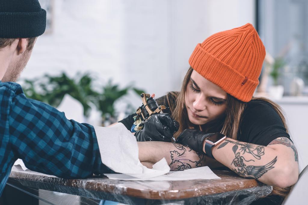 Молодой человек и женщина-татуировщик во время процесса татуировки в студии
 - Фото, изображение