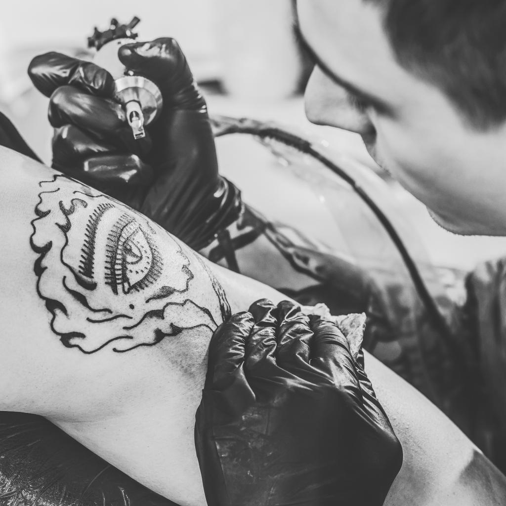 Μαύρο και άσπρο φωτογραφία του καλλιτέχνη τατουάζ στα γάντια εργασίας σε ώμο κομμάτι στο studio - Φωτογραφία, εικόνα