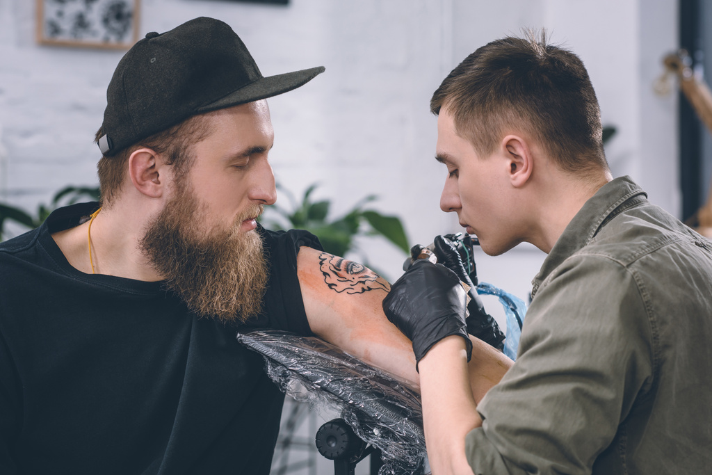 Artista del tatuaje y hombre barbudo durante el proceso de tatuaje en el estudio
 - Foto, Imagen