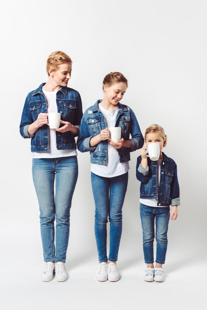 mother and daughters in similar denim clothing with cups of hot drinks standing in row isolated on grey - Photo, Image