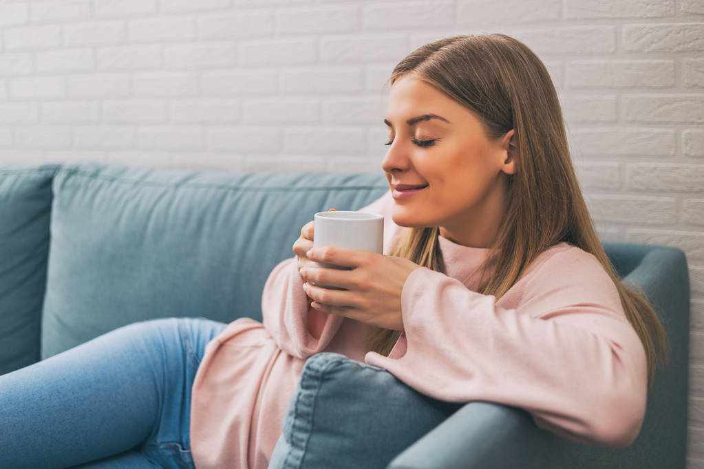 Piękna kobieta cieszy, picie kawy, podczas odpoczynku na kanapie w swoim domu. Obraz jest celowo stonowanych. - Zdjęcie, obraz