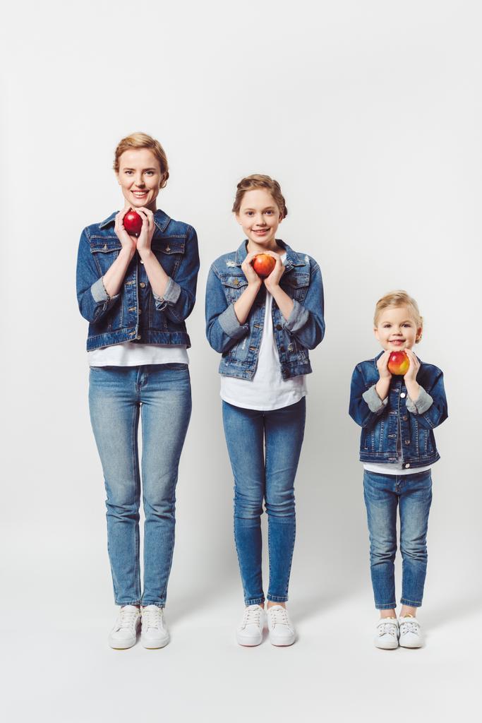 lachende moeder en dochters van verschillende generaties in soortgelijke kleding met verse appels geïsoleerd op grijs - Foto, afbeelding