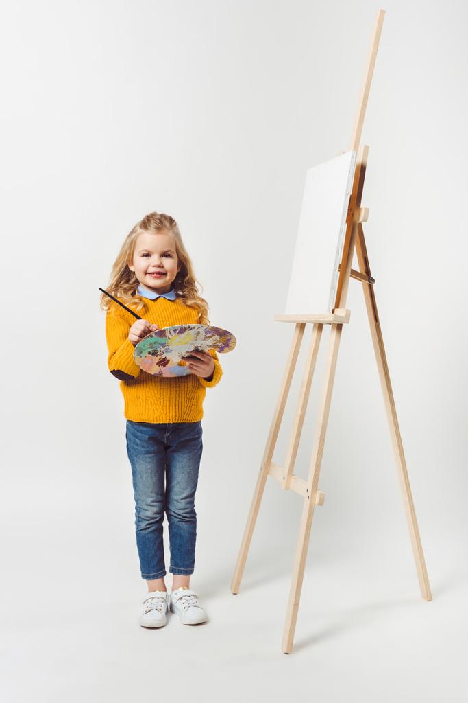 suloinen pieni lapsi, jossa on sivellin ja paletti seisoo lähellä kangasta maalaustelineessä valkoisella
 - Valokuva, kuva