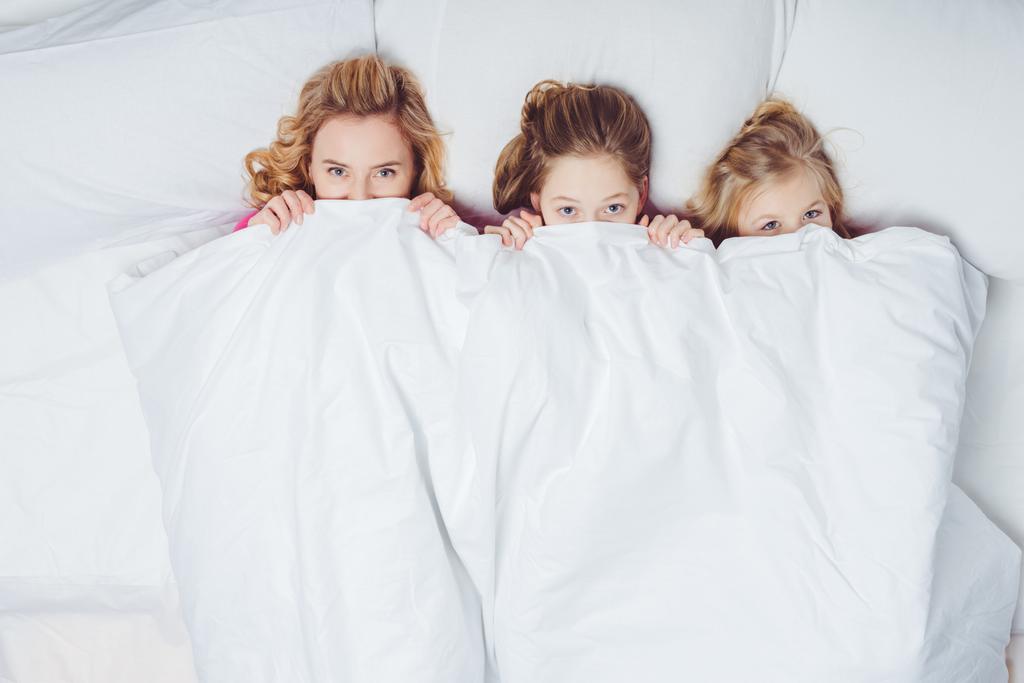 top view of mother and daughters hiding under blanket in bed - Photo, Image