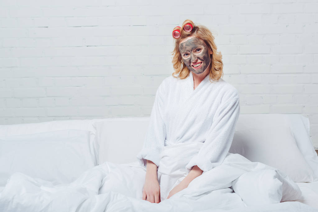 mooie vrouw in badjas met gezichtsmasker zitten in bed - Foto, afbeelding