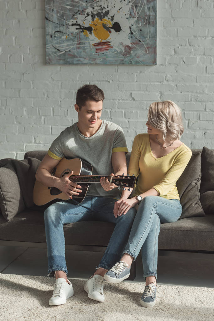 knappe vriendje spelen van akoestische gitaar voor vriendin thuis - Foto, afbeelding