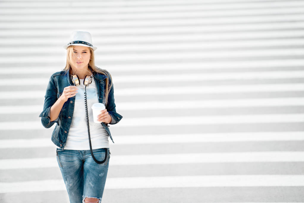 Kaunis nuori nainen hattu ja farkut takki takeaway kahvikuppi, seisoo tiellä seepra rajan yli, juo kahvia, ja poseeraa tiellä tausta
. - Valokuva, kuva