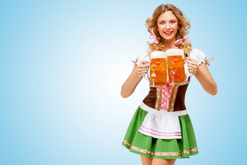 jonge sexy oktoberfest vrouw dragen een traditionele Beierse jurk dirndl dienen twee bier mokken met happy glimlach op blauwe achtergrond. - Foto, afbeelding
