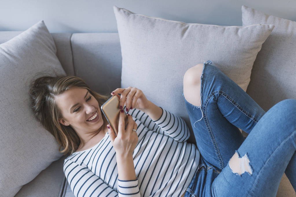 Девушка читает сообщение на смартфоне. Счастливая женщина использует мобильный телефон на диване. Счастливая женщина дома читает смс-ку в своей светлой гостиной. Расслабленная молодая женщина смс сообщения на диване
 - Фото, изображение