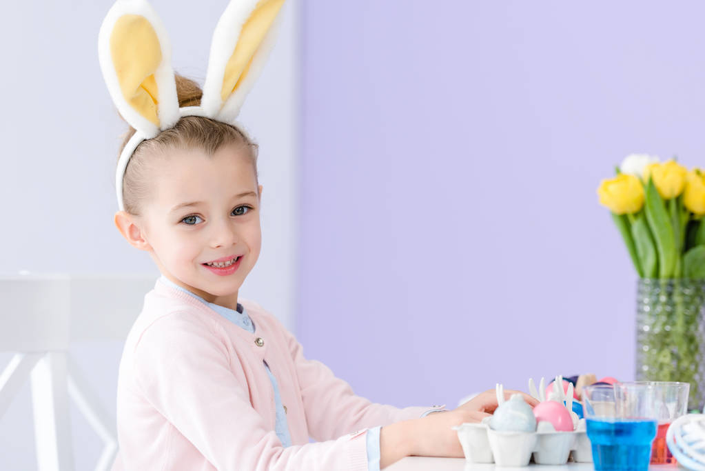 Kind meisje spelen bunny oren dragen door tafel met Pasen gekleurde eieren - Foto, afbeelding