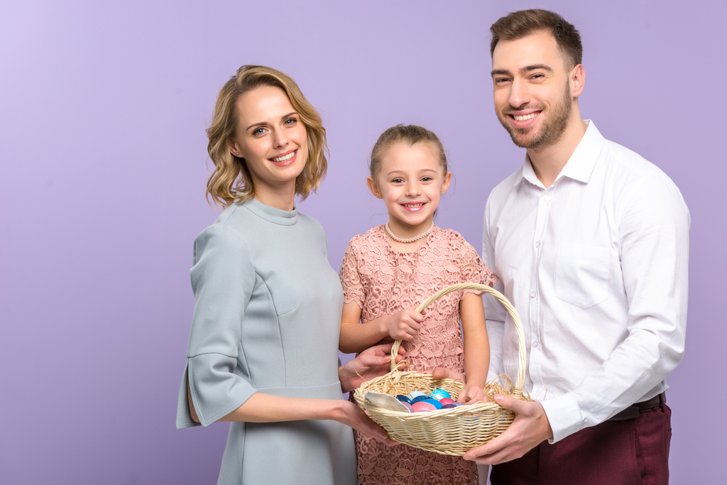 Padres e hija con cesta de Pascua con huevos de colores aislados en violeta
 - Foto, imagen