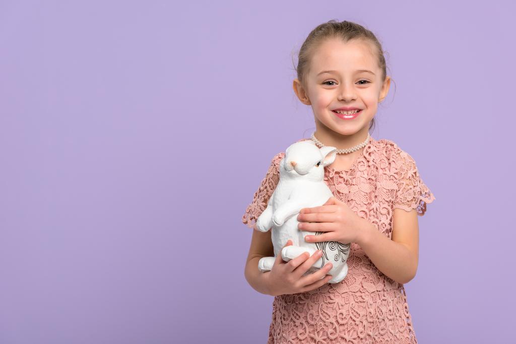 kleines Mädchen mit Hasenstatuette zu Ostern isoliert auf violett - Foto, Bild