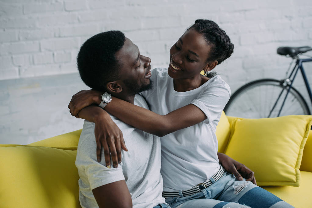 feliz jovem casal afro-americano abraçando enquanto sentado no sofá em casa
 - Foto, Imagem