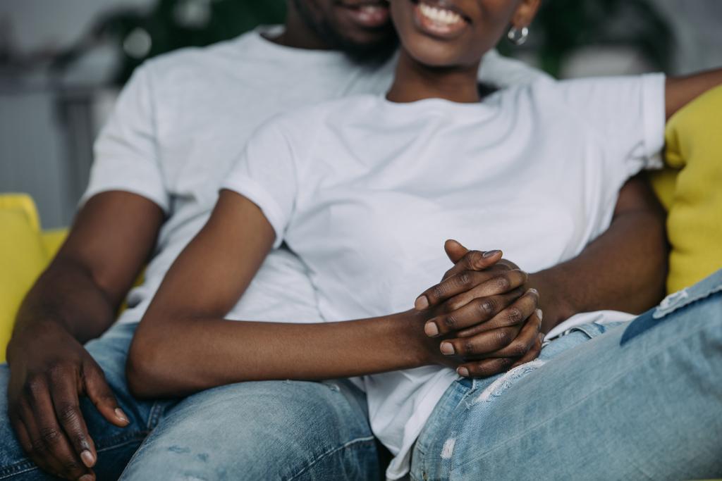 oříznutý snímek mladý americký pár v bílých tričkách drží ruce, zatímco sedí doma - Fotografie, Obrázek