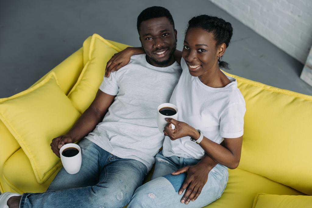 hoge hoekmening van jonge Afrikaanse Amerikaanse echtpaar met kopjes koffie zittend op de Bank en glimlachend op camera - Foto, afbeelding