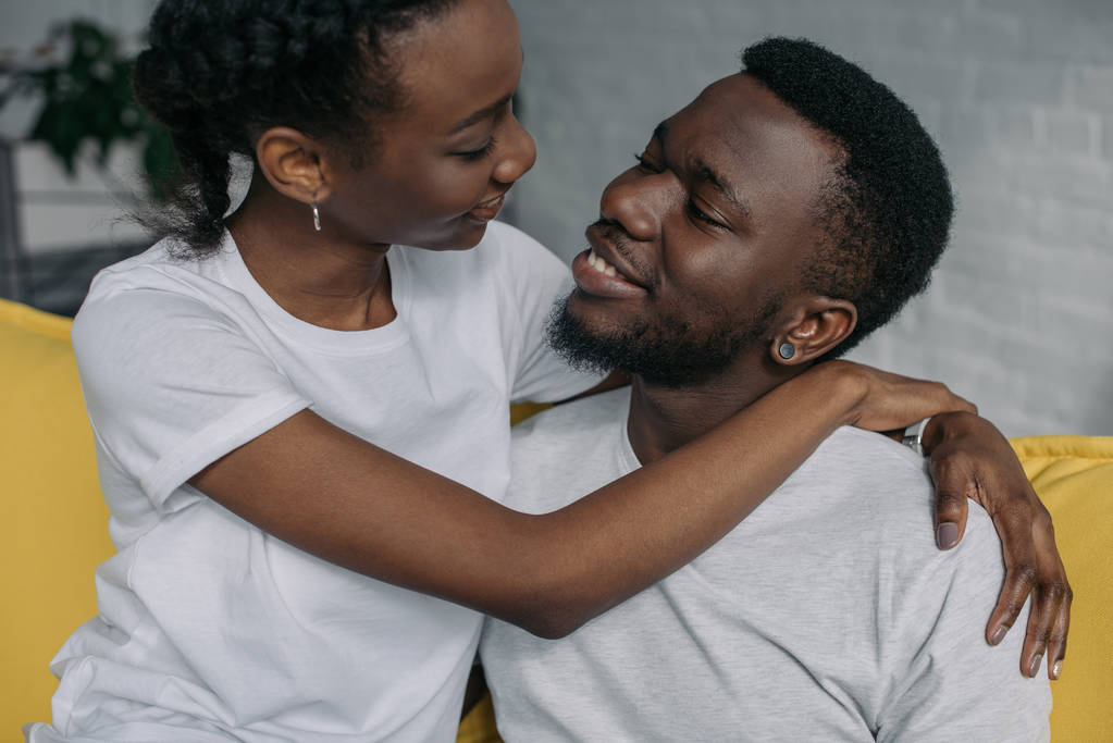 young african american couple in white t-shirts hugging and smiling each other at home - Photo, Image
