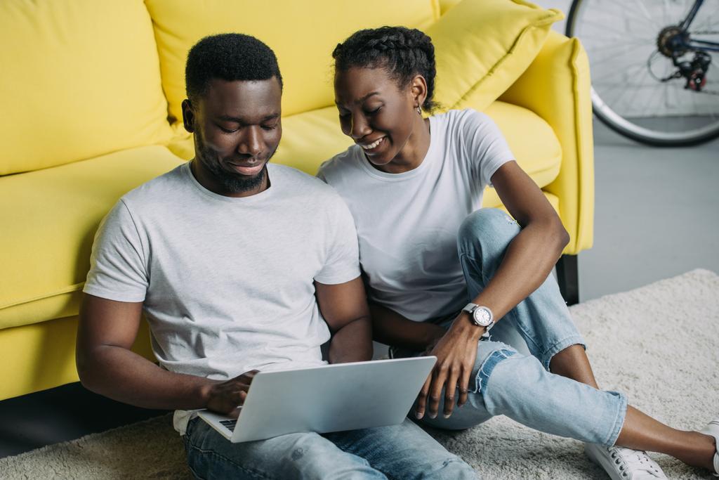 χαρούμενος νεαρός αφρικανική αμερικανική ζευγάρι σε λευκά μπλουζάκια χρησιμοποιώντας φορητό υπολογιστή μαζί στο σπίτι - Φωτογραφία, εικόνα