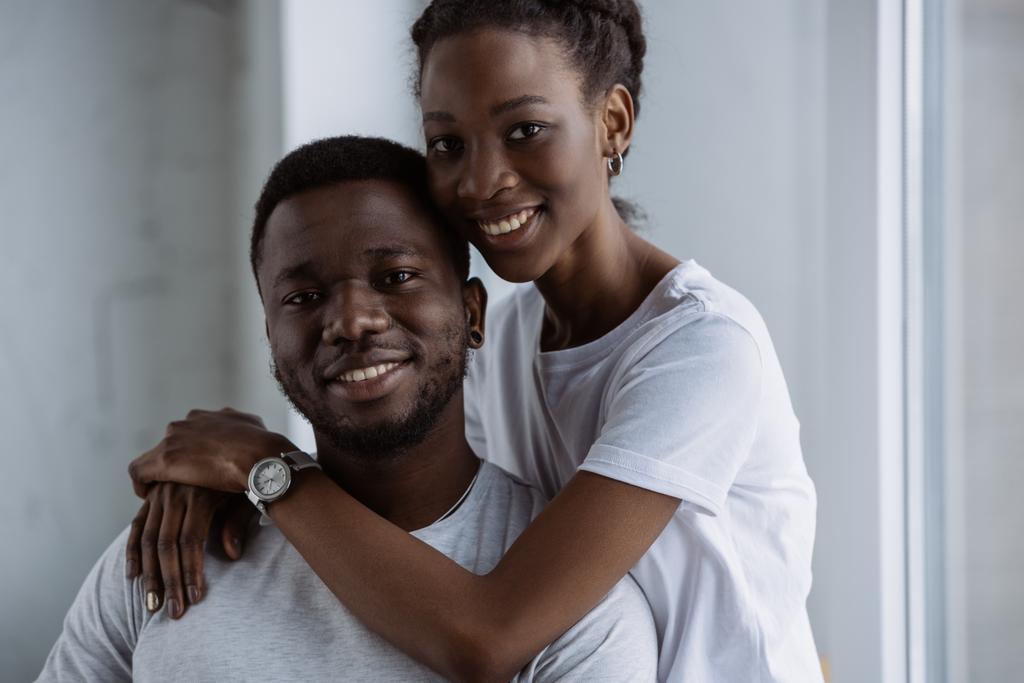 счастливая молодая африканская американская пара в белых футболках обнимающая и улыбающаяся в камеру
  - Фото, изображение