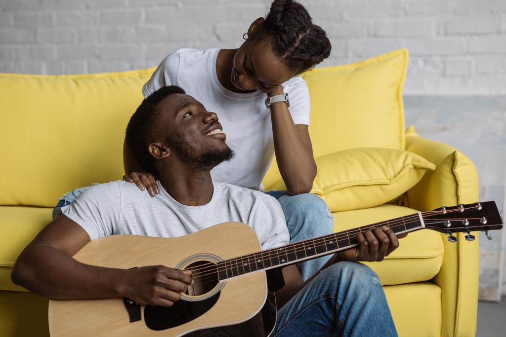 schöner lächelnder junger Mann, der Gitarre spielt und die schöne Freundin zu Hause ansieht - Foto, Bild