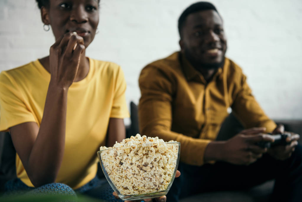 oříznuté záběr africké americké dívky jíst popcorn zatímco přítel hraje s joystickem doma - Fotografie, Obrázek