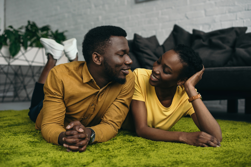 mooie jonge Afrikaanse Amerikaanse echtpaar liggen op tapijt en glimlachen elkaar thuis - Foto, afbeelding