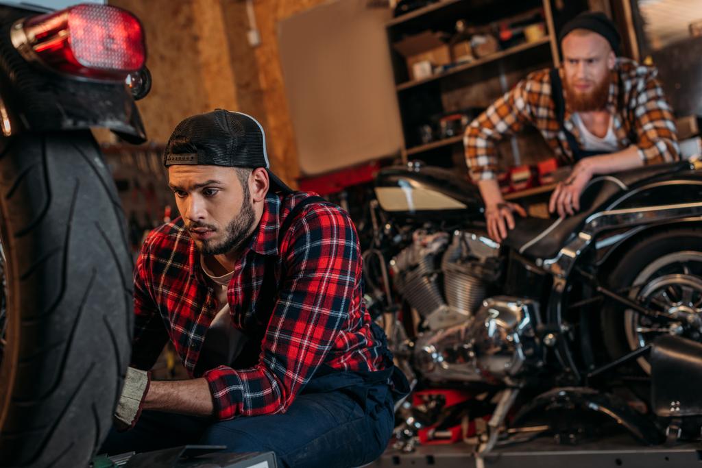 сфокусированный механик чинит мотоцикл, пока его коллега стоит на заднем плане
 - Фото, изображение