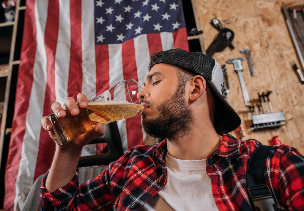 trabajador de la estación de reparación guapo beber cerveza en el garaje con bandera EE.UU. colgando en la pared
 - Foto, Imagen
