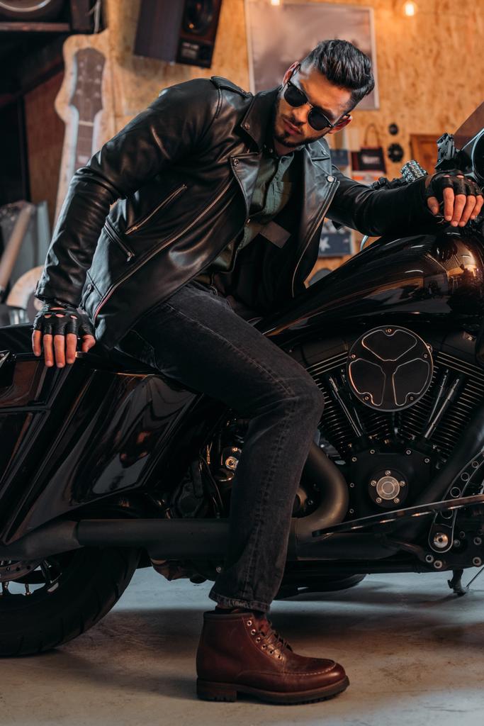 pohledný mladý muž ve stylovém oblečení sedí na motocyklu v garáži - Fotografie, Obrázek