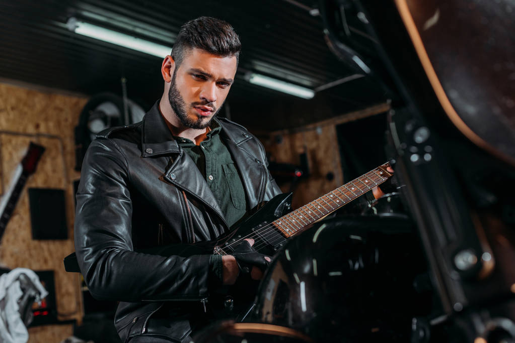 joven guapo tocando la guitarra mientras está sentado en bicicleta en el garaje
 - Foto, Imagen