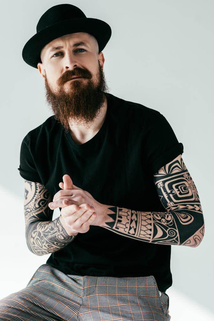 όμορφος γενειοφόρος άνδρας τατουάζ στο καπέλο κοιτάζοντας την κάμερα στο λευκό - Φωτογραφία, εικόνα