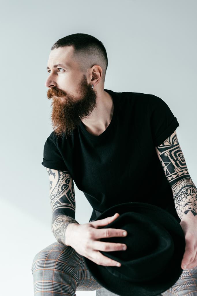 όμορφος γενειοφόρος τατουάζ άνθρωπος κρατάει καπέλο και αναζητούν μακριά σε λευκό - Φωτογραφία, εικόνα