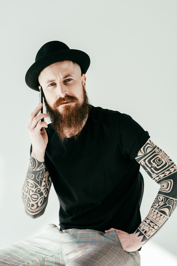 parrakas tatuoitu mies puhuu älypuhelimella ja katselee kameraa valkoisella
 - Valokuva, kuva