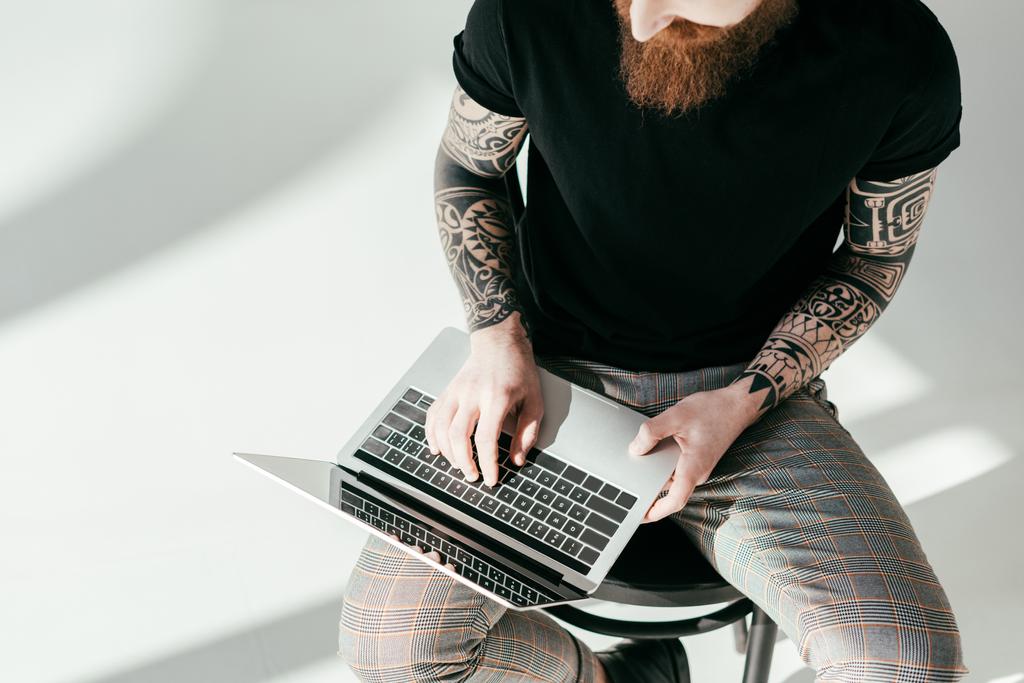 обрезанное изображение бородатого татуированного мужчины, сидящего на стуле с ноутбуком на белом
 - Фото, изображение