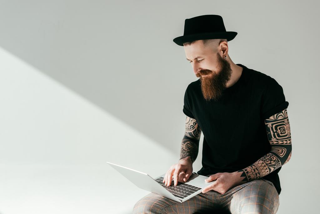 όμορφος γενειοφόρος άνδρας τατουάζ κοιτάζοντας το laptop στο λευκό - Φωτογραφία, εικόνα