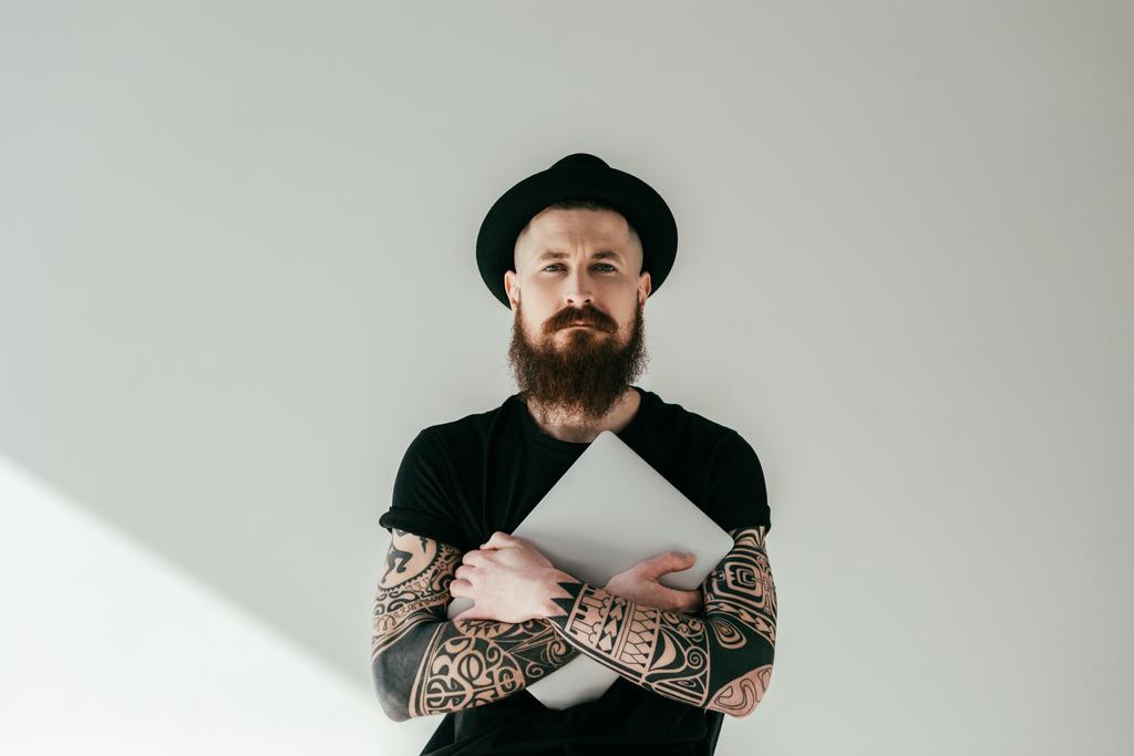 guapo barbudo hombre tatuado abrazando portátil y mirando a la cámara en blanco
 - Foto, Imagen