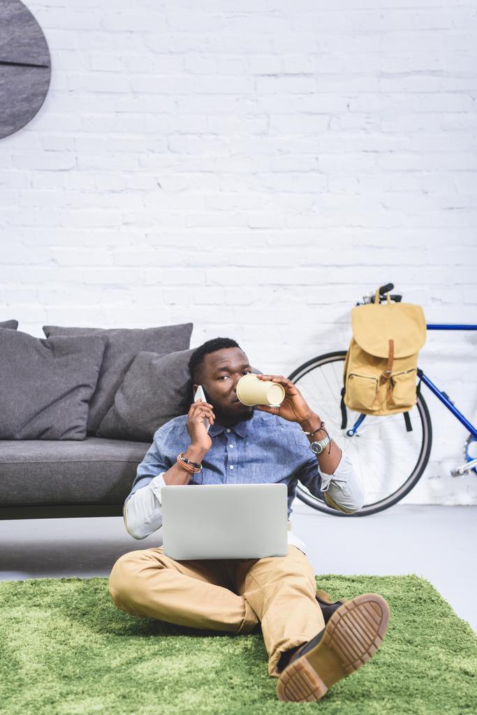 Όμορφος αφρικανική αμερικανική άνθρωπος μιλώντας στο τηλέφωνο και πίνοντας καφέ ενώ εργάζονται για το laptop - Φωτογραφία, εικόνα